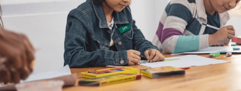 Students Drawing at a Table, One Girl Wears a Name Tag Labeled "Jane"
